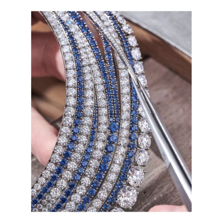 Close-up of ornate jewelry construction with white and blue gemstones, held by hands and adjusted with tweezers, on a wood surface.