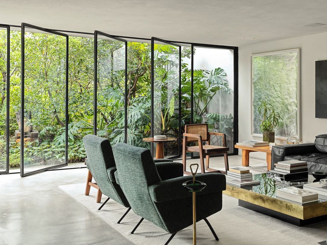 Modern interior living room featuring green armchairs, a glass coffee table, and a view of lush greenery through folding glass doors.