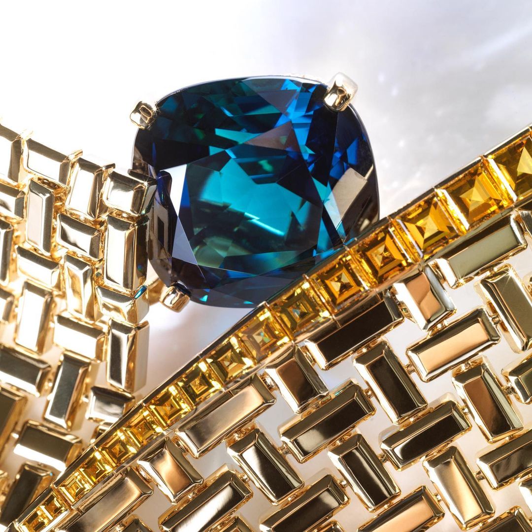 Close-up of a blue gemstone surrounded by a gold, rectangular-patterned setting on a white background, fashion jewelry product shot.