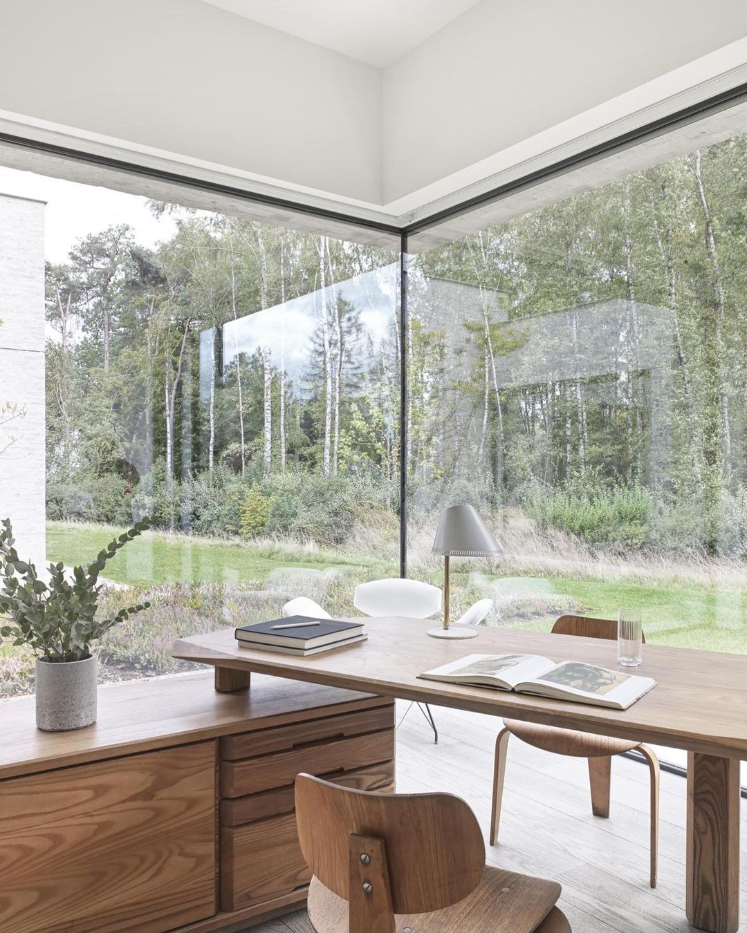 Wooden desk and chair in a room with large windows overlooking a forest, with a view of a modern house in the background.