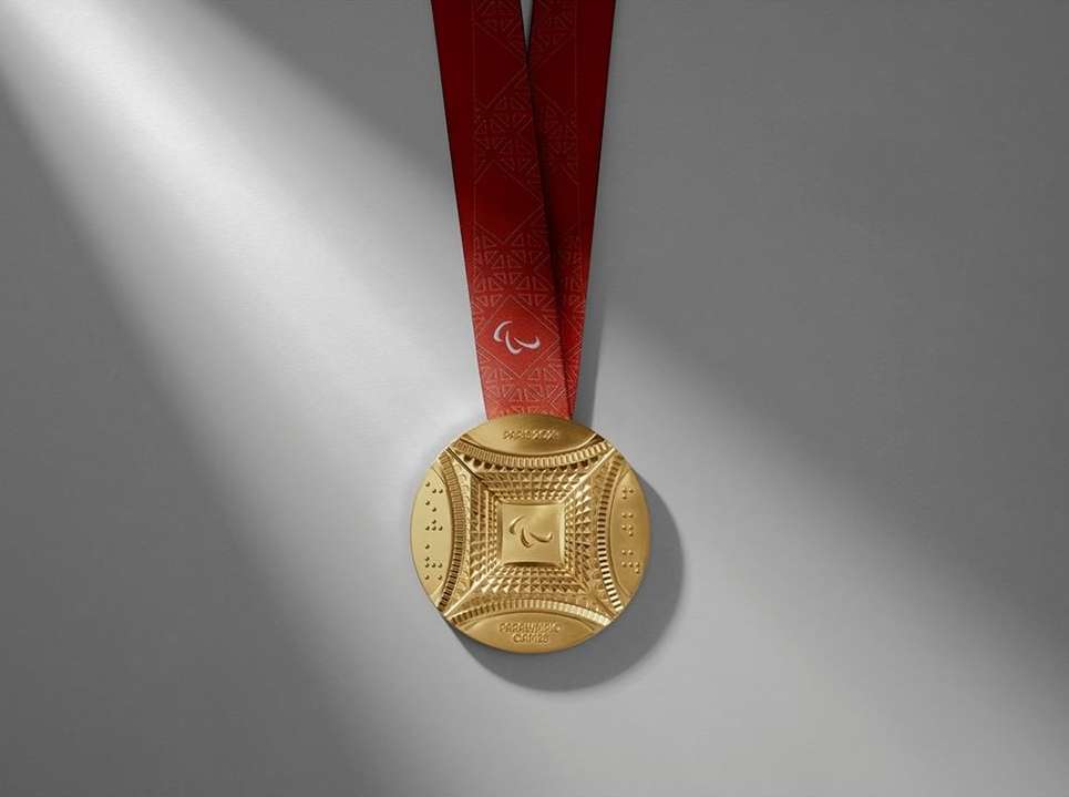 Close-up of a gold-colored Paralympics medal hanging from a red patterned ribbon, casting a soft shadow on a neutral grey background.
