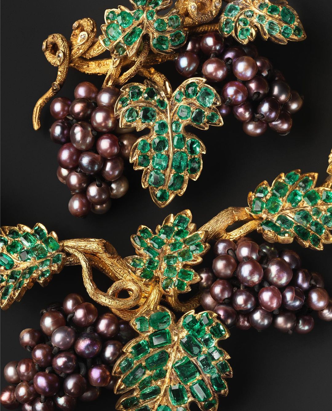 Close-up of a jewelry artwork featuring gold vine structures, emerald-like leaves, and pearl-like purple grapes on a black background.