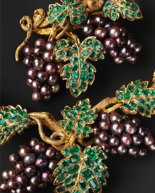Close-up of a jewelry artwork featuring gold vine structures, emerald-like leaves, and pearl-like purple grapes on a black background.