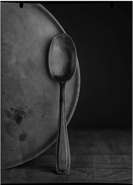 Monochrome still life of a metal spoon resting against a plate on a wooden surface, captured in a high-contrast, textured black and white photograph.