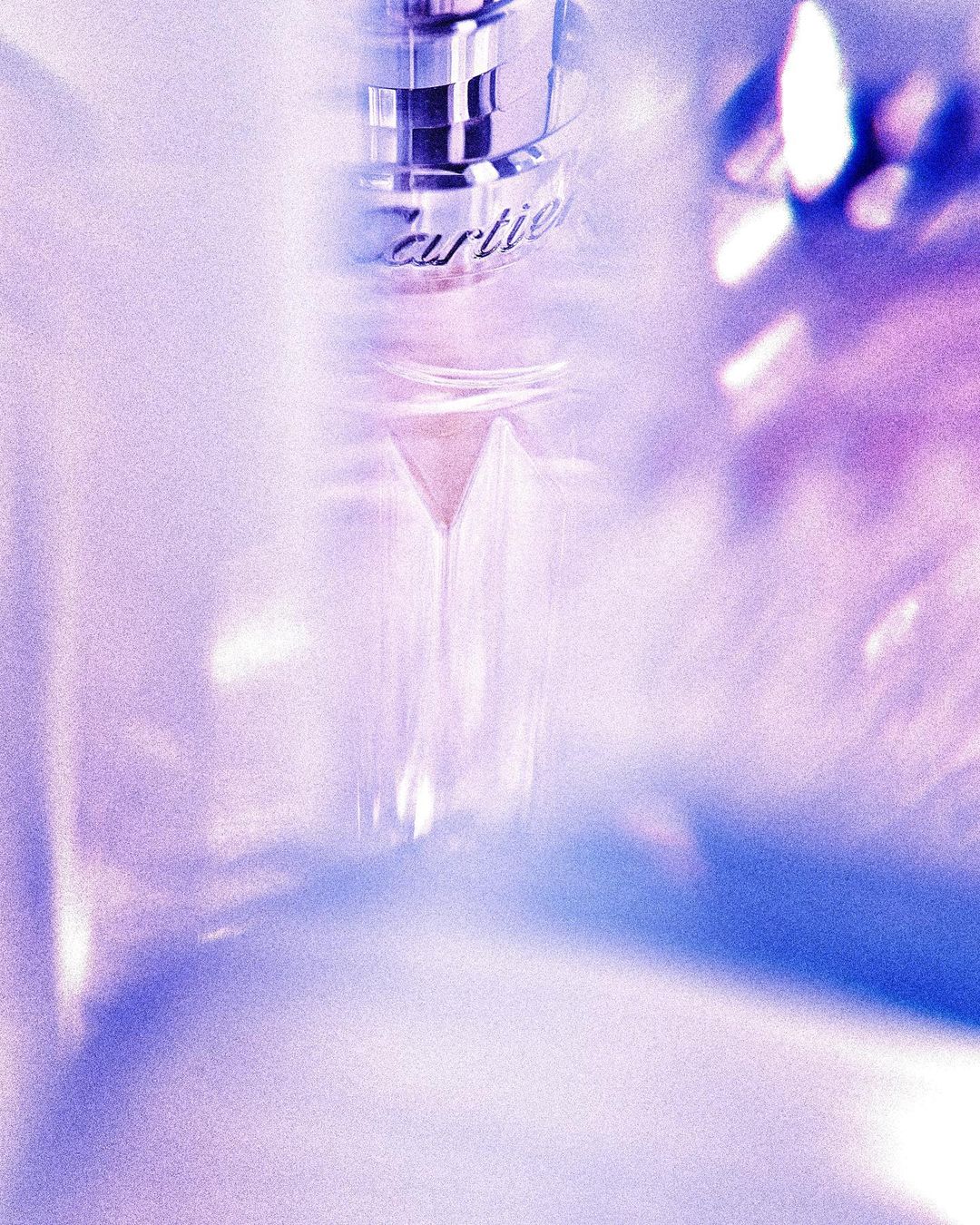 Close-up product shot of a clear glass perfume bottle with metallic silver accents, against a soft, light purple backdrop.