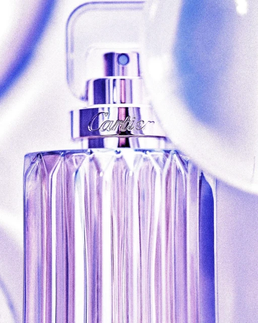 Close-up of a faceted glass perfume bottle with silver accents against a soft, blurred background. The 'Cartier' logo is visible.