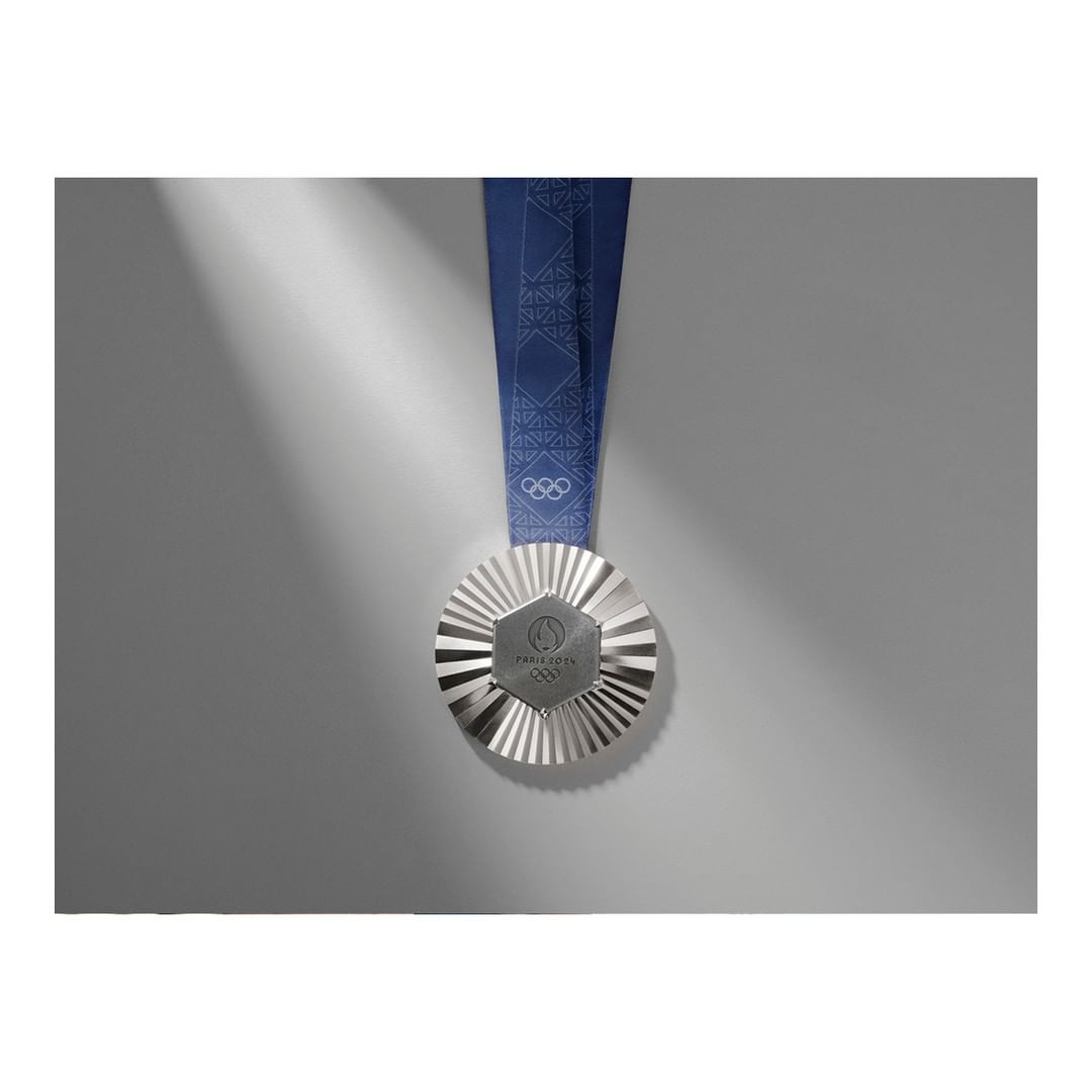 Close-up of a silver medal with a blue ribbon hanging on a light gray background with a beam of light shining on the left side.