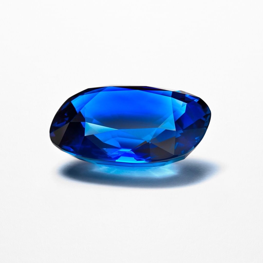 Close-up of a faceted, oval-cut bright blue gemstone on a white surface, reflecting light and shadow. Jewel for jewelry design.