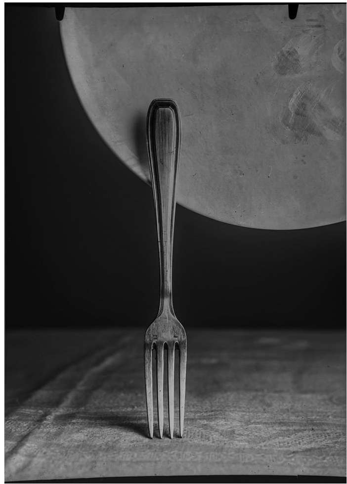 Monochrome photo of a silver fork standing upright on a textured surface, with a partial round plate in the dark background.