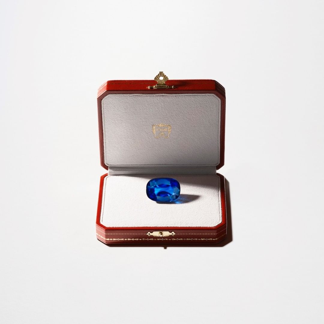 Close-up of a vibrant blue gemstone inside an open red and cream jewelry box, centered against a bright white background.