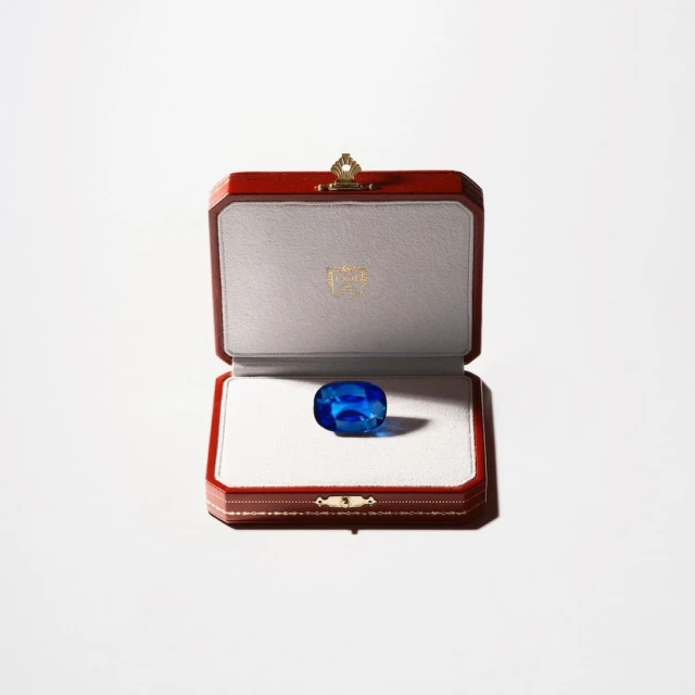 Close-up of a vibrant blue gemstone inside an open red and cream jewelry box, centered against a bright white background.