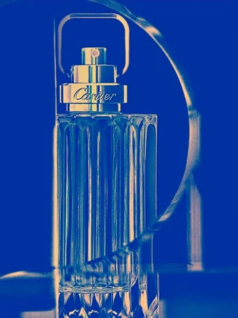 A clear glass Cartier perfume bottle with a gold collar is set against a striking blue background, with a metallic ribbon accent.