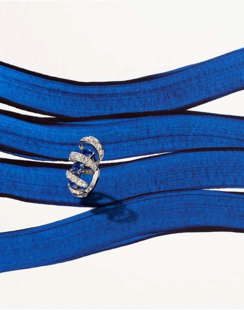 Statement jewelry shot. A solitaire ring with blue gemstone and diamond spirals lays on strips of blue and white fabric.