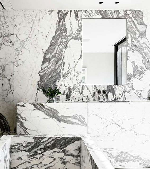 Modern bathroom features grey and white marble walls and floors, a rectangular mirror, a glass vase, and minimalist design elements.