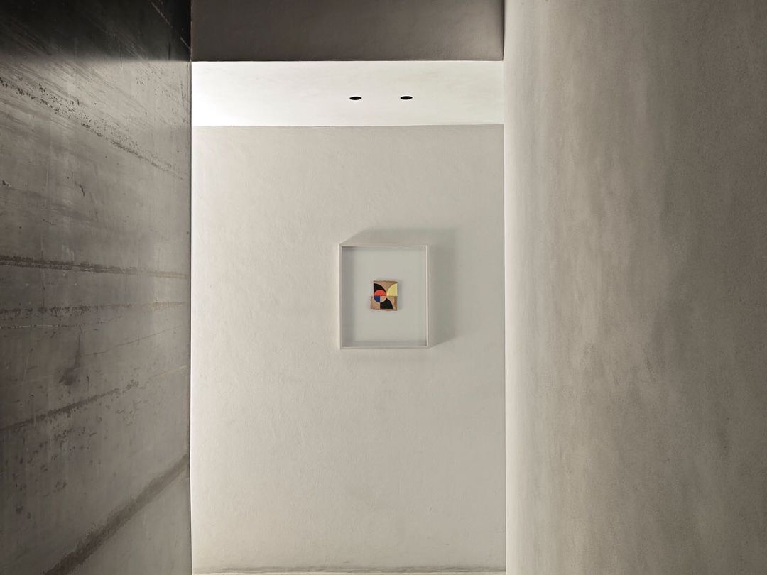 Modern interior corridor with concrete walls, leading to a white wall featuring an abstract painting in a white frame.