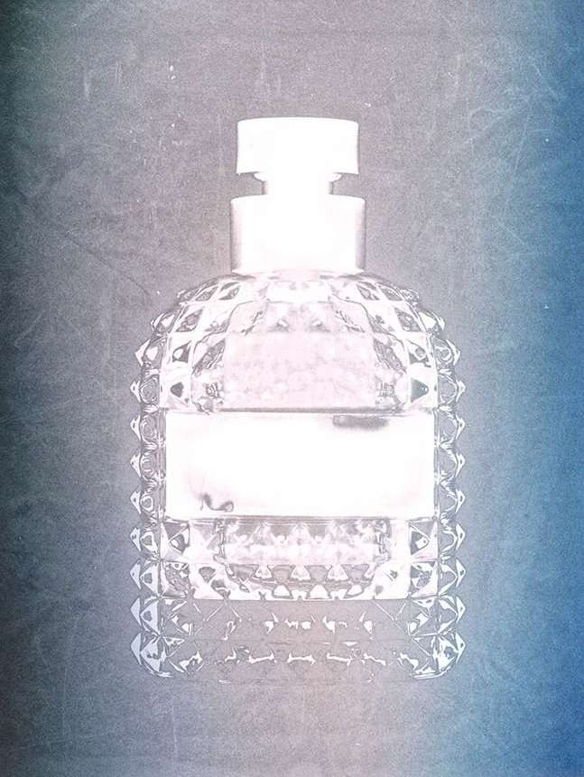 Close-up of a clear, faceted glass perfume bottle with a white cap and label against a textured blue and grey gradient background.