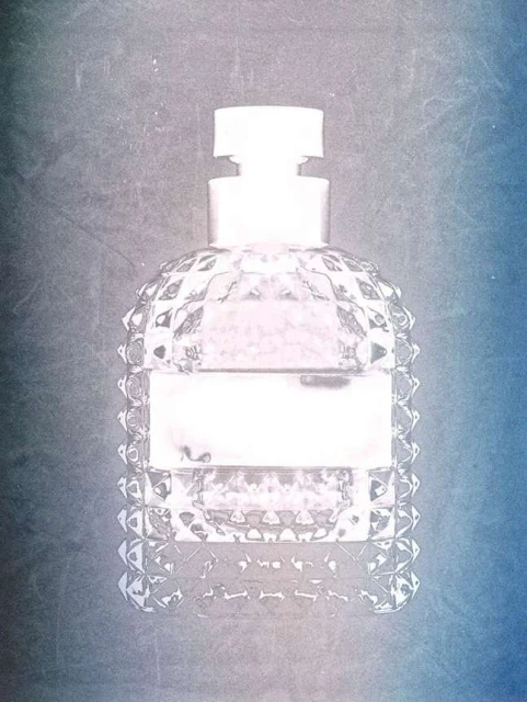 Close-up of a clear, faceted glass perfume bottle with a white cap and label against a textured blue and grey gradient background.