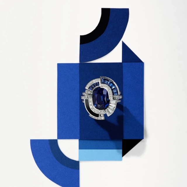 High-fashion ring with large blue gemstone surrounded by diamonds, resting on geometric paper art in shades of blue and white.