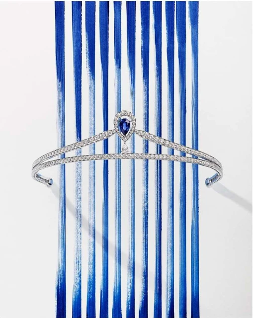 Elegant tiara with a sapphire teardrop accent, set against a backdrop of vibrant blue brushstrokes on a white surface, showcasing luxury and artistry.