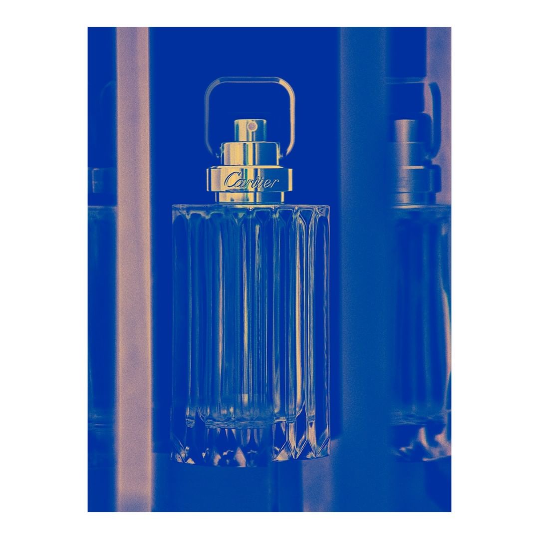A clear glass perfume bottle with a metal handle against a vibrant blue duotone background by Thomas Deschamps.
