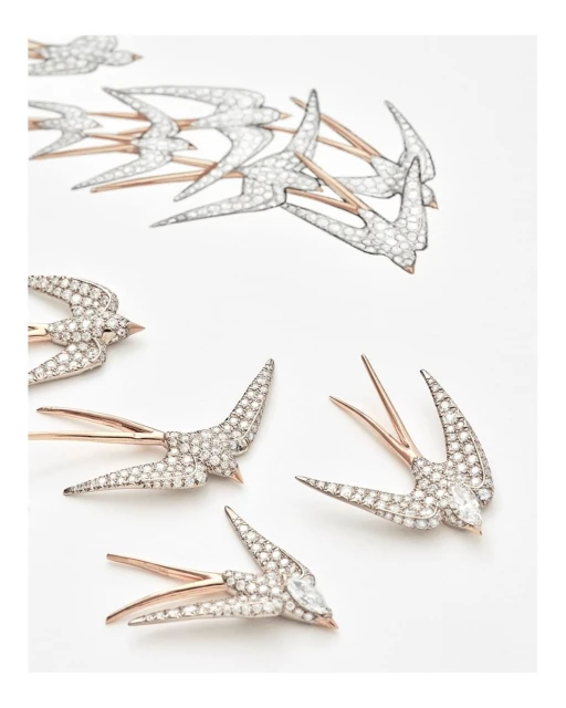 Close-up of several swallow-shaped brooches with silver bodies and gold wings scattered on a white surface, studio lighting.