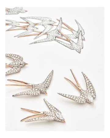 Close-up of several swallow-shaped brooches with silver bodies and gold wings scattered on a white surface, studio lighting.