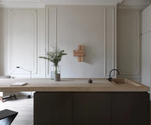 Minimalist kitchen interior featuring a long stone island, dark cabinetry, and ornate molding on the pale wall.
