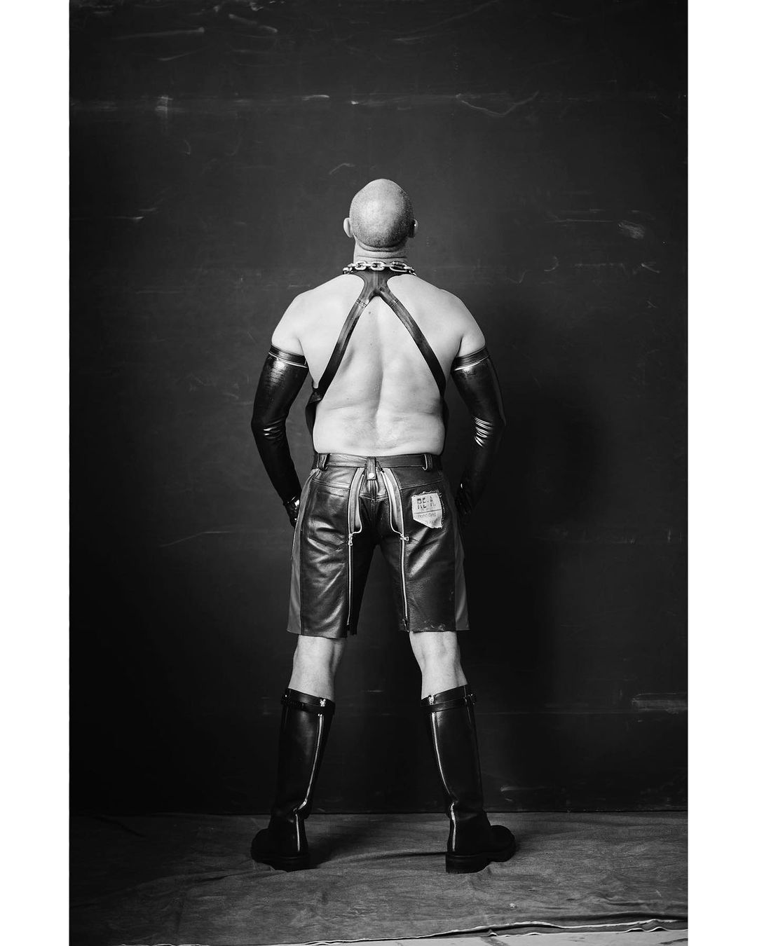Black and white shot of a bald, muscular adult male from the rear, wearing leather shorts, gloves, and boots, against a dark wall.