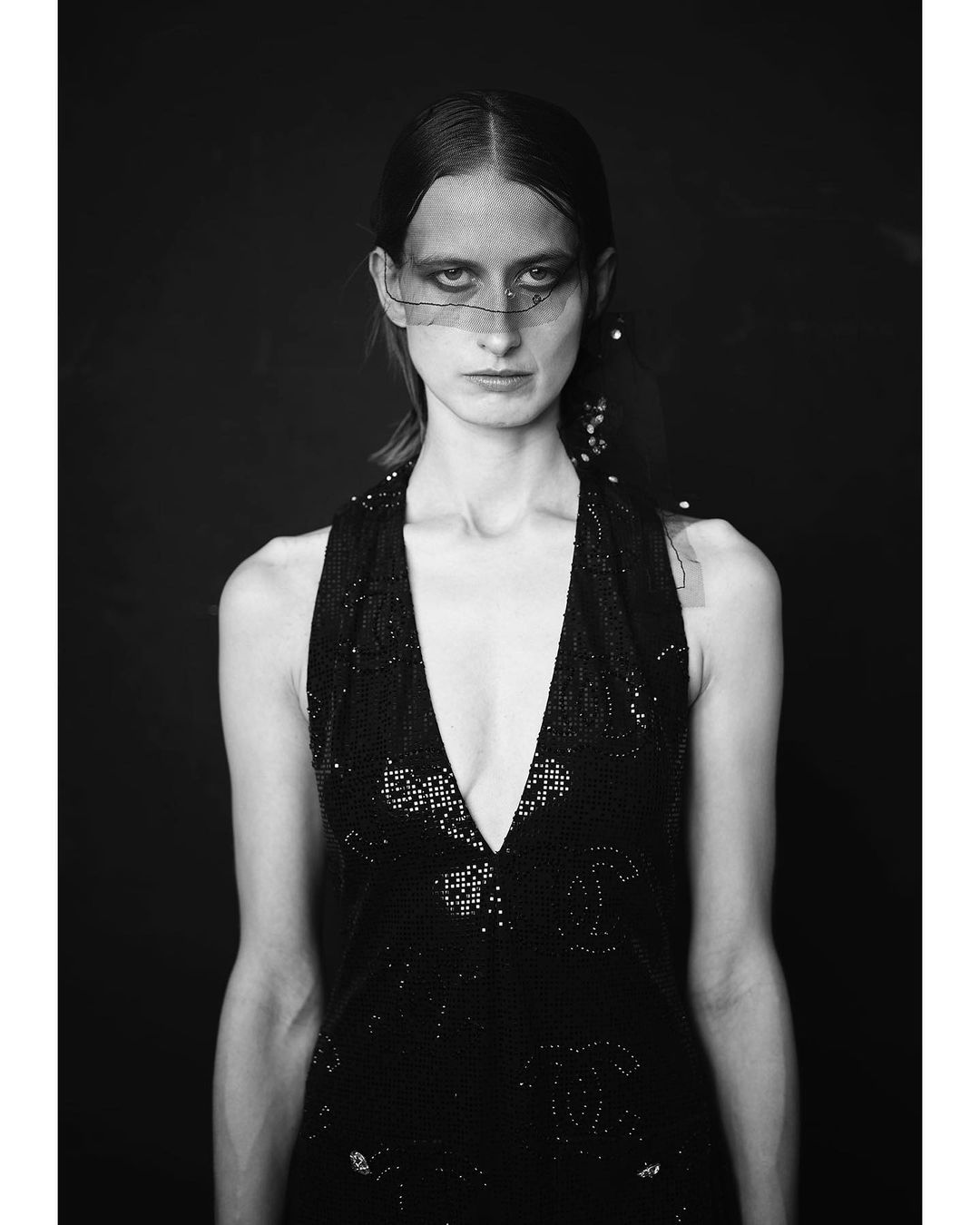 Black and white portrait of a model in a glittering halter dress and veiled headpiece against a dark background in a studio.