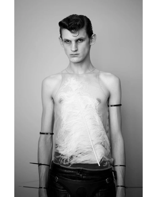 Black and white portrait of a young adult man with slicked back hair, wearing a sheer top, and leather pants posing in a photo studio.