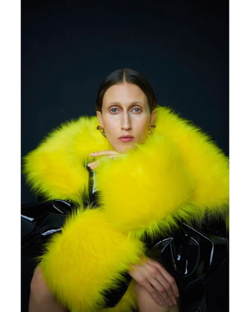 Fashion portrait of a woman with dark hair wearing a fluffy yellow jacket and black patent leather outfit against a dark background.