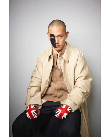 Man with black paint on his face wearing a beige coat, brown cardigan, and Union Jack gloves against a grey background.