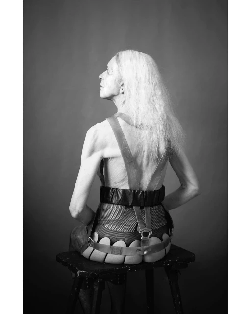 Back view of elderly female sitting on stool with brace around torso, long white hair against grey background in a black-and-white studio shot.