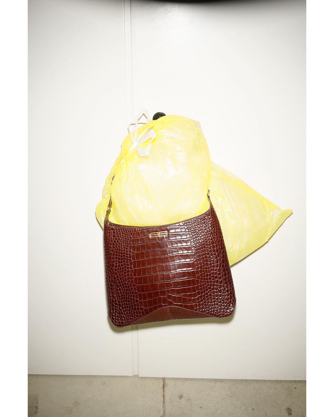 Brown faux-crocodile purse hangs in front of a partially visible yellow plastic bag against a white wall indoors.