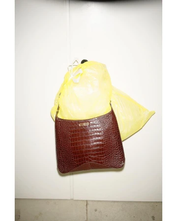 Brown faux-crocodile purse hangs in front of a partially visible yellow plastic bag against a white wall indoors.