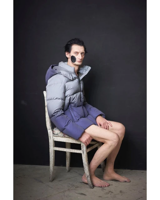 Man with black face paint sits on white chair wearing puffy ombre down jacket against black background in studio setting.
