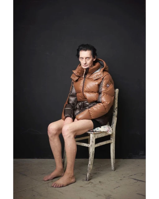 A person with bare legs sits on a chair wearing a puffy brown hooded jacket in a studio setting with a dark background.