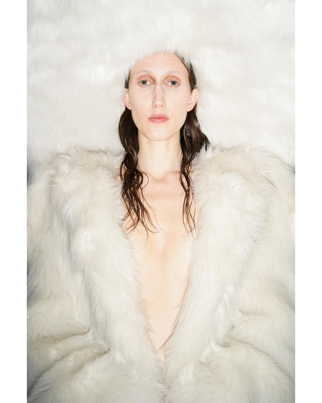 Fashion shot of a woman with brown hair wearing a big fur coat and fuzzy hat looking at the camera in a brightly lit indoor space.