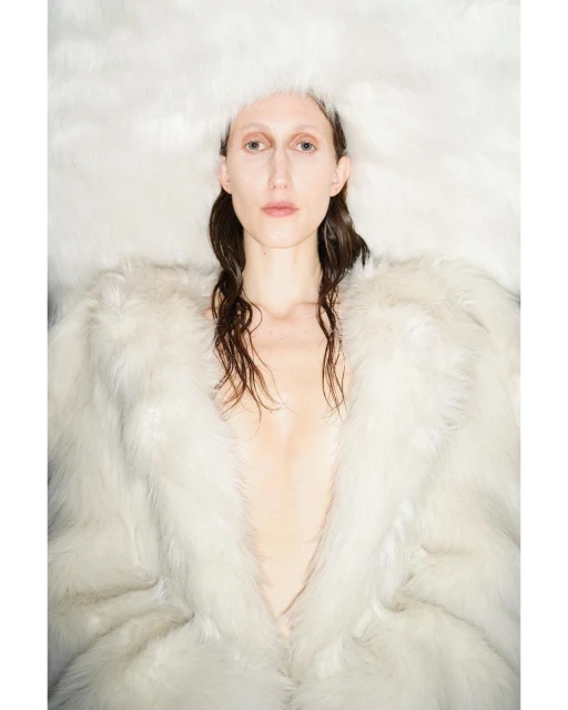 Fashion shot of a woman with brown hair wearing a big fur coat and fuzzy hat looking at the camera in a brightly lit indoor space.