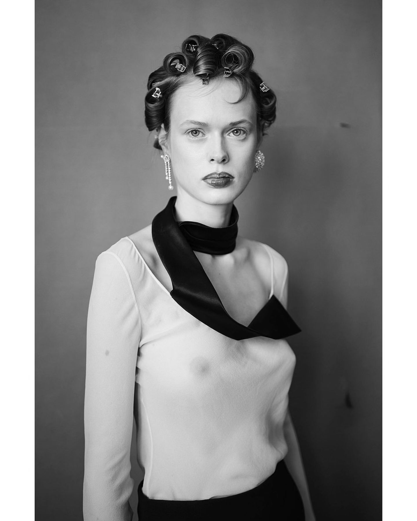Monochrome portrait of a woman with curlers, sheer top, and black neck scarf against a grey studio backdrop, capturing a retro aesthetic.