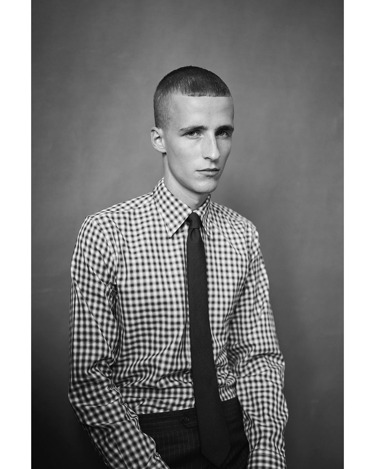 Black and white portrait of a young adult male wearing a checkered shirt and tie, gazing directly at the camera against a plain backdrop.