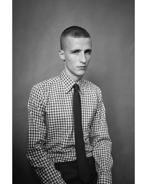Black and white portrait of a young adult male wearing a checkered shirt and tie, gazing directly at the camera against a plain backdrop.