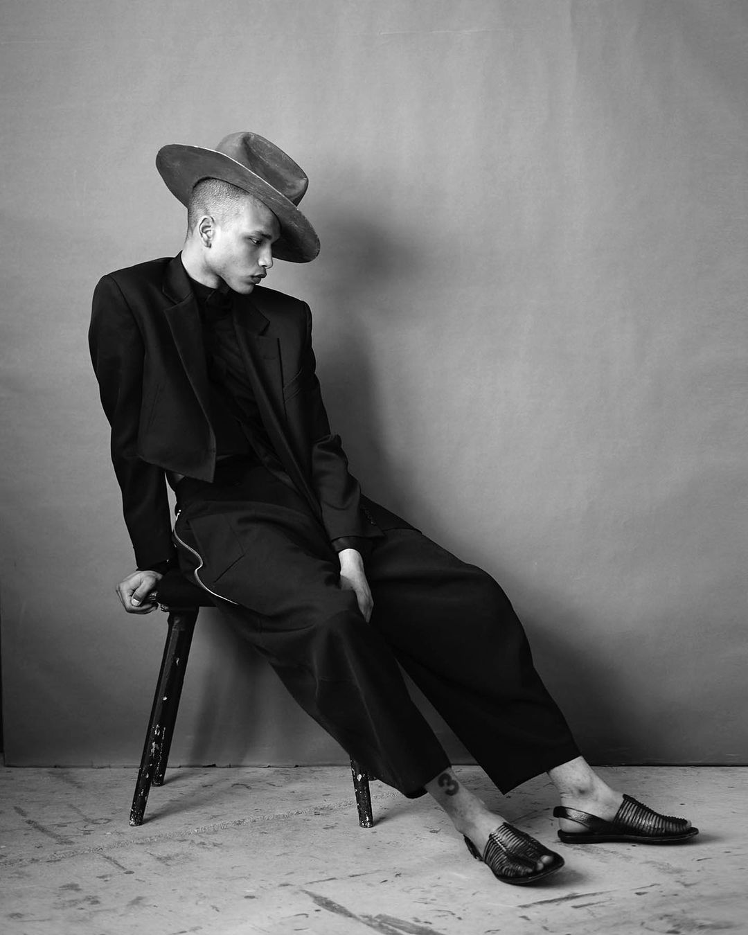 Black and white portrait of a young adult in a black suit and hat, seated on a stool against a neutral background in a studio setting.