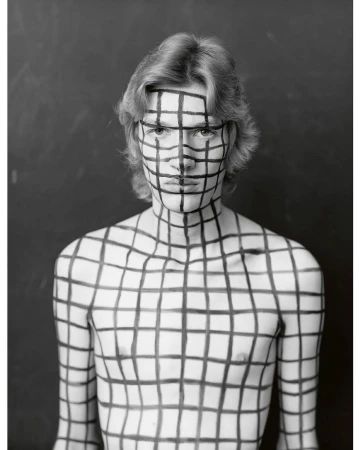 Portrait of a man with bold black grid pattern painted across his face and bare upper body against dark background.