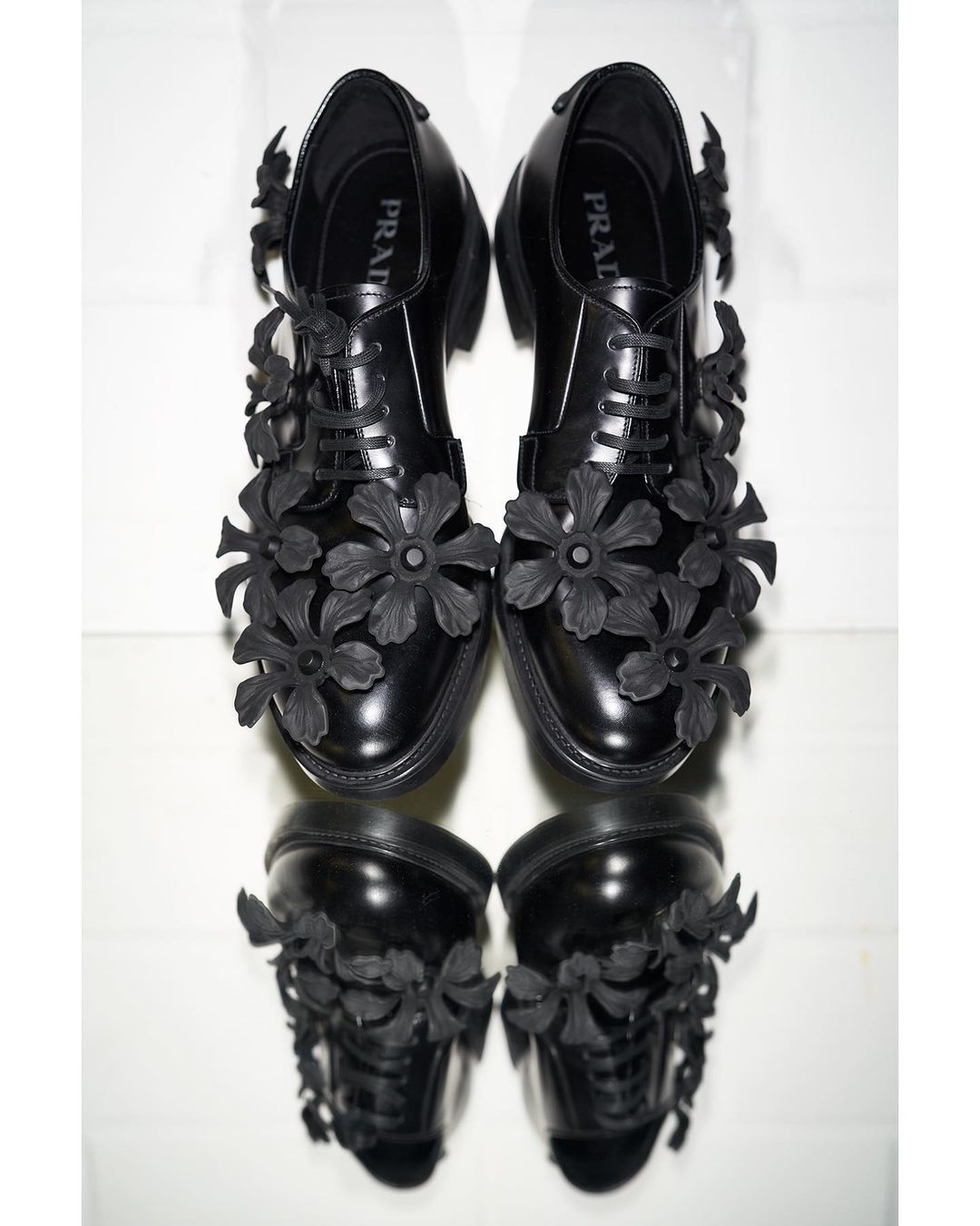 A pair of black leather Prada dress shoes decorated with floral accents. The shoes are on a white surface with a mirrored reflection.