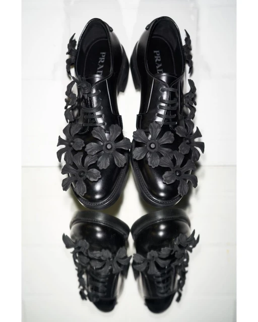 A pair of black leather Prada dress shoes decorated with floral accents. The shoes are on a white surface with a mirrored reflection.