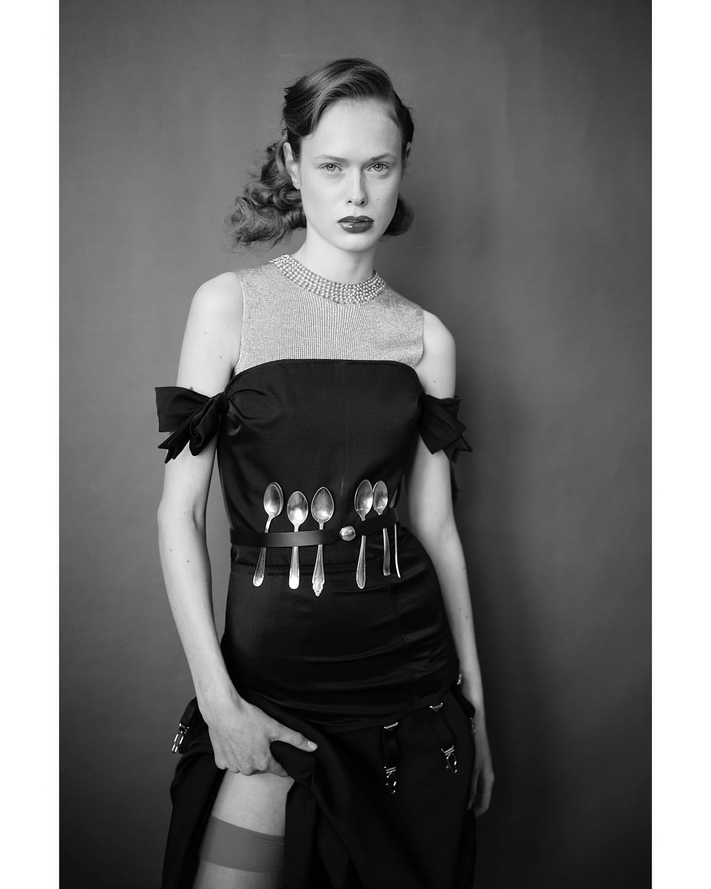 Black and white portrait of a female fashion model in a black dress adorned with spoons, standing in front of a grey background.