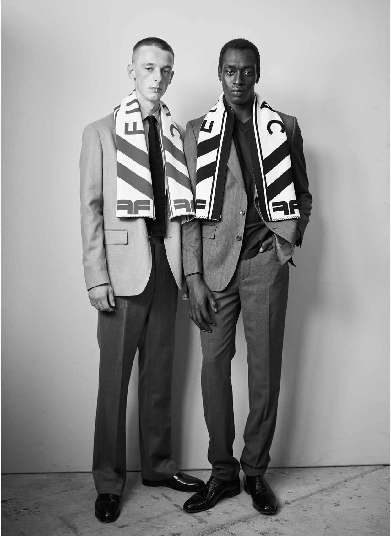 Two men in suits and scarves stand indoors. Man on the left is light-skinned, the man on the right is dark-skinned. Black and white photo.