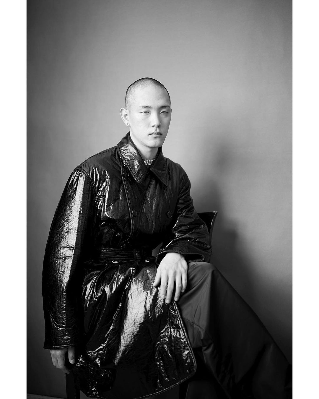 Monochrome studio portrait of a bald adult male seated wearing a shiny black vinyl trench coat with a gray background and a serious expression.