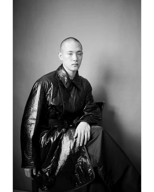 Monochrome studio portrait of a bald adult male seated wearing a shiny black vinyl trench coat with a gray background and a serious expression.